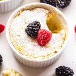 White ramekin with a lemon pudding cake in it. Topped with a raspberry and blackberry. A scoop of the cake is scooped out and on a spoon next to the ramekin.