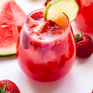 A glass of Strawberry Watermelon Rosé Sangria with a round lime slice on the edge of the glass. Watermelon slices and strawberries surrounding the glass.