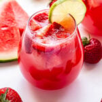 A glass of Strawberry Watermelon Rosé Sangria with a round lime slice on the edge of the glass. Watermelon slices and strawberries surrounding the glass.