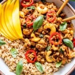 Closeup overhead photo of Thai Basil Chicken on top of rice in a white bowl with chopsticks. Garnished with mango slices, sliced Thai chiles and basil leaves.
