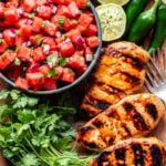Overhead photo of grilled honey lime chicken with a bowl of watermelon salsa, squeezed lime halves and jalapaños behind the chicken. A bunch of cilantro next to the chicken.