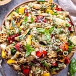 Overhead photo of Serving bowl filled with grilled zucchini, summer squash, tomatoes, corn and pasta. Coated in pesto and garnished with basil, parmesan cheese and pine nuts. Small bowl of pesto and a chunk of parmesan cheese behind the bowl.