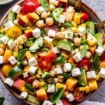 Overhead photo of chickpea, cucumber, tomato, peach, feta, basil salad in serving bowl with spoons in the bowl. A small bowl of lemon vinaigrette behind the salad.