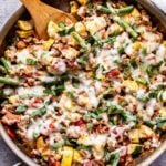 Overhead photo of Ground turkey, zucchini, summer squash, green beans and tomatoes topped with melted mozzarella cheese in metal skillet with a wooden spoon in the skillet.