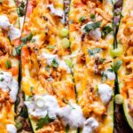 Overhead photo of sheet pan with two zucchini halves filled with buffalo chicken, topped with melted cheese, ranch dressing, green onions and cilantro.
