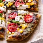 Overhead photo of a corner slice of Grilled Corn, Tomato and Pesto Pizza with dollops of ricotta and basil leaves.
