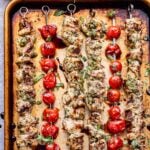 Overhead photo of four grilled pesto chicken skewers and two grilled tomato skewers on a sheet pan. Basil leaves and a bowl of pesto behind the sheet pan.