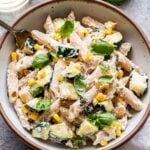 Overhead photo of a white bowl filled with penne pasta, zucchini, corn, basil leaves and a creamy ricotta sauce. There is a fork in the bowl and a glass of white wine behind the bowl.