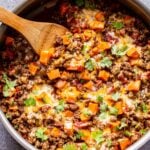 Overhead photo of a metal skillet filled with ground beef, tomatoes, sweet potatoes, southwest spices and topped with melted cheese and cilantro. Wooden serving spoon in the skillet.