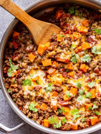 Overhead photo of a metal skillet filled with ground beef, tomatoes, sweet potatoes, southwest spices and topped with melted cheese and cilantro. Wooden serving spoon in the skillet.