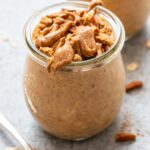 Pumpkin pie overnight oats in a jar topped with graham cracker crumbs, pecans, and almond butter. A spoon is in front of the jar.