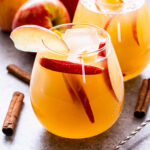 Front view of two glasses filled with apple cider sangria. Apple slices are in the cocktails and the rims of the glasses have an apple slice for garnish. Apples and cinnamon sticks are surrounding the glasses.