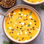 Overhead photo of A white bowl with a spoon in it, filled with creamy chipotle sweet potato soup topped with a drizzle of Greek yogurt, cilantro and pepitas. A small wooden bowl is behind the soup and filled with pepitas.