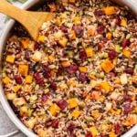 Wild rice stuffing with butternut squash, apples, cranberries, pecans and pepitas in a metal skillet with a wooden spoon in it.