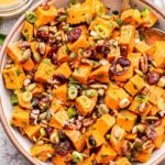 Maple Orange Sweet Potato Salad in a white serving bowl with spoons. A jar of the vinaigrette behind the bowl.