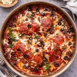 Baked pizza chicken topped with melted cheese, pepperoni and black olives in a metal skillet. Bowl of shredded cheese behind the skillet.