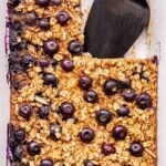 Blueberry Maple Baked Oatmeal in a white baking dish with a corner piece cut out and a black spatula in it's place.