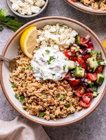 Greek chicken bowl with rice, tzatziki and tomato cucumber salad in a white bowl with a small bowl of feta cheese behind it.