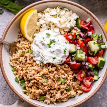 Greek chicken bowl with rice, tzatziki and tomato cucumber salad in a white bowl with a small bowl of feta cheese behind it.