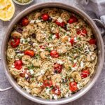 grilled shrimp pesto pasta with feta, olives, and tomatoes in a stainless steel skillet with a bowl of pesto and a lemon behind the skillet..
