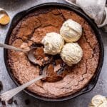 Mexican Skillet Brownies with two spoons in the skillet and three scoops of vanilla ice cream on top of them. An ice cream scoop with vanilla ice cream in it in front of the skillet.