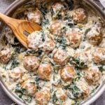 spinach artichoke dip meatballs in a metal skillet with a wooden spoon in the skillet.