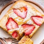 A slice of strawberry sheet pan pancake topped with syrup on a white plate with a fork holding a piece of the edge.