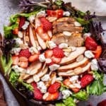 Grilled Chicken Salad with Strawberries and Goat Cheese in a bowl with wooden serving spoons to the left and a bowl of balsamic vinaigrette behind it.