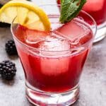 blackberry gin fizz cocktail in a glass with a lemon slice and mint sprig for garnish. A bottle of gin and a second cocktail are behind the glass.