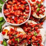 white serving plate with grilled chicken breasts topped with melted mozzarella and tomato basil topping. A bowl of the tomato basil topping on the plate as well.