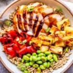 White bowl filled with teriyaki chicken, pineapple, red pepper, edamame and brown rice.