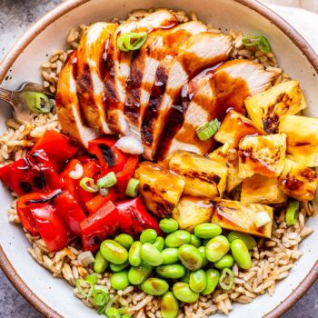 White bowl filled with teriyaki chicken, pineapple, red pepper, edamame and brown rice.