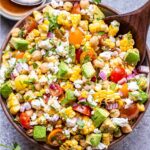 Tomato Avocado Corn Salad in white serving bowl with a wooden spoon in it. A bowl of vinaigrette and half of a lime are behind the salad bowl.