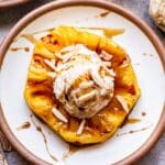 A white plate with a grilled pineapple ring topped with vanilla ice cream and rum sauce.