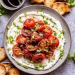Grilled Tomatoes with Ricotta topped with fresh herbs and balsamic glaze on a gray plate. Slices of baguette next to the plate and a bowl of balsamic glaze behind it.