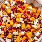Roasted butternut squash with goat cheese and pomegranate seeds in a white bowl with spoons and a jar of balsamic vinaigrette behind it.