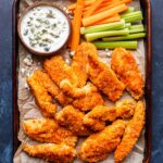 parchment paper lined sheet pan with Baked Buffalo Chicken Tenders, a bowl of blue cheese dip, and carrot and celery sticks.