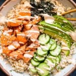White bowl filled with rice and topped with baked salmon, sliced cucumber and avocado, nori, and spicy mayo.