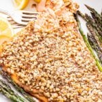 Walnut Crusted Salmon on a white platter with asparagus and lemon wedges. The edge is being flaked with a fork.