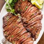 Two sliced carne asada flank steaks on a white platter with lime wedges and cilantro.
