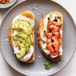 Two slices of Burrata Toast on a gray plate. One topped with strawberries and the other with avocado slices.