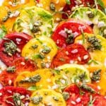 Red, yellow, green and orange sliced Heirloom Tomatoes on a white plate topped with pesto, pine nuts, and basil.