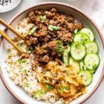 Beef Bulgogi on top of rice in a white bowl with chopsticks. Also topped with kimchi and sliced cucumbers.