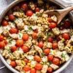 Pesto Chicken Tomato and Corn in a stainless steel pan with a wooden serving spoon in it. A bowl of pesto is behind the pan.
