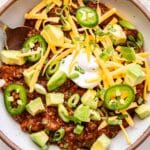 Overhead photo of a white bowl with no bean chili in it. Topped with cheese, avocado, scallions, and Greek yogurt.