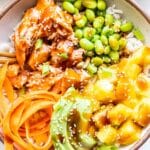 Teriyaki Salmon rice Bowl in a white bowl. Topped with edamame, pineapple, avocado slices, and carrot ribbons.