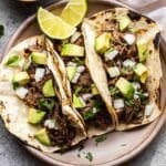Three barbacoa beef tacos on a plate with lime wedges.