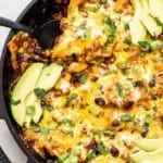 Baked ground beef enchilada skillet topped with melted cheese and avocado slices. A black serving spoon is scooping some out.