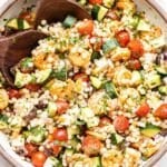 Mediterranean Couscous Salad with shrimp in a white serving bowl with wooden serving spoons.