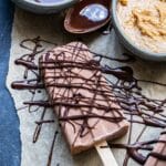 Chocolate Peanut Butter Banana Yogurt Pop drizzled with chocolate with a bowl of peanut butter and melted chocolate behind it.